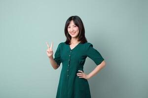 Southeast asian woman smiling confidently in green dress posing with peace sign for advertising on plain background with positive energy photo