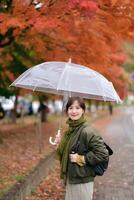 Southeast asian woman stands smiling under transparent umbrella on rainy autumn day with vibrant orange leaves creating warm and peaceful atmosphere in background photo