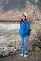 Southeast asian woman enjoys travel concept standing near river with bridge in background wearing blue jacket and white beanie expressing happiness in nature photo