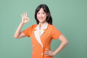 Southeast asian woman in orange uniform smiling and showing ok hand gesture on green background expressing positivity photo