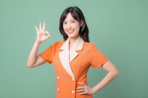 Southeast Asian woman in orange uniform smiling and making OK hand gesture stands confidently against green background showing positive and cheerful emotion photo