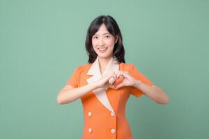 Southeast asian woman in orange dress smiling and making heart gesture with hands on green background expressing happiness photo