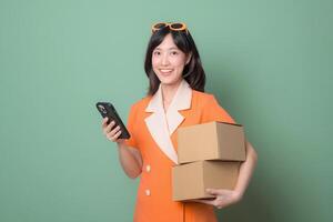 Southeast asian woman holding packages and using smartphone for internet online shopping, smiling and wearing orange dress with sunglasses against green background photo