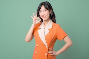 Southeast Asian woman in orange dress smiling and making OK hand gesture stands confidently against green background showing positive and cheerful emotion photo