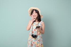 Southeast asian woman in summer dress holding phone and camera, wearing straw hat, smiling with joyful travel expression against pastel background photo