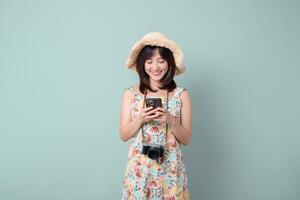 Happy Asian woman in summer dress and straw hat holding phone and camera, smiling on pastel green background photo