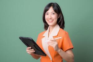 Southeast asian woman smiling while using internet on digital tablet with stylus pen, standing against green background, professional and confident expression photo