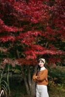 Southeast asian woman stands smiling under vibrant red autumn leaves wearing brown sweater and white pants enjoying peaceful outdoor nature scene in fall season photo