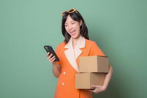 Southeast asian woman in orange dress holding cardboard box and using smartphone with happy expression on green background photo