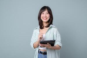 Southeast Asian woman smiling while using internet on digital tablet, casual outfit, standing against plain background, cheerful and confident expression, modern lifestyle photo