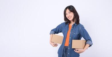 Southeast asian woman in denim shirt smiling and holding two cardboard boxes against white background, showing cheerful and confident expression suitable for delivery or moving concept photo