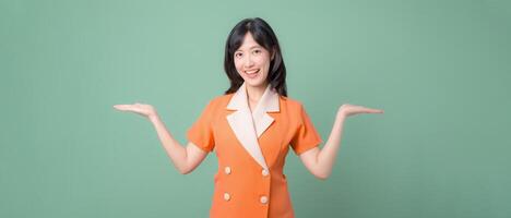 Southeast asian woman in orange suit smiling with arms raised on green background expressing cheerful confidence photo