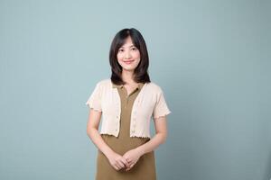 Southeast Asian woman with medium length black hair stands smiling in front of plain blue background wearing short sleeve beige cardigan and olive dress, expressing calm confidence photo