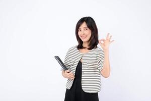 Southeast Asian woman smiling and holding tablet while making OK hand gesture, standing against white background, expressing confidence and positivity photo