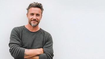 A man with a beard and grey sweater standing in front of a white wall photo