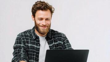 A man with a beard and a plaid shirt is smiling while using a laptop photo