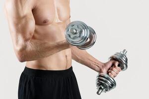 Unrecognizable muscular bodybuilder with dumbbells pumping up muscles over light studio background, copy space. Determined personal coach exercising with weights, working out his biceps photo