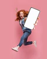 Excited redhead woman having fun, jumping,demonstrating mobile phone with empty white screen on pink studio background. Creative mockup collage with space for website or app design photo