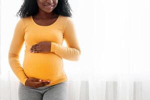 Smiling black lady carrying baby, hugging her big tummy while standing next to window at home. Cheerful african american pregnant woman embracing her belly and smiling at camera, happy to be mother photo