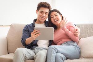 Chinese Couple Making Call Via Digital Tablet, Communicating Remotely Online Sitting On Couch At Home, Waving Hands To Computer Screen. Distance Communication Concept. Copy Space photo