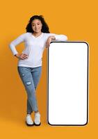 Cool Mobile App. Cheerful Black Woman Leaning At Big Smartphone With Blank White Screen, Demonstrating Copy Space For Your Design Or Advertisement, Standing Over Yellow Background, Mockup Image photo