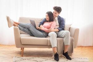 Chinese Boyfriend And Girlfriend With Laptop Watching Movie Online Relaxing On Weekend Sitting On Sofa At Home. Couple Spending Time Together, Using Computer And Browsing Internet. Empty Space photo