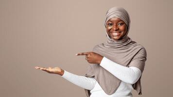 Smiling Black Muslim Lady In Hijab Pointing Finger At Her Open Palm, Demonstrating And Advertising Something, Posing Over Light Studio Background, Holding Invisible Object And Smiling, Copy Space photo