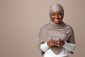 Modern Of Smiling Black Islamic Woman In Hijab Using Smartphone And Smiling At Camera, Downloading New Application, Browsing Internet Or Texting With Friends, , Posing Over Light Studio Background photo