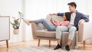 Asian Couple Using Smartphone Mobile Application Relaxing Sitting On Sofa In Minimalistic Living Room At Home On Weekend. Phone Users Browsing Internet. Gadget Lifestyle, Mobile Apps. Copy Space photo