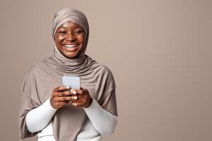 Joyful Black Muslim Lady In Hijab Holding Smartphone And Laughing At Camera, Enjoying Mobile Communication And Online Shopping, Browsing New App, Standing On Light Studio Background, Copy Space photo