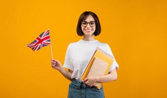 Travel and study, learn English language. Zoomer smiling woman with notebooks and small flag go to university, college or school, isolated on orange background, studio shot photo