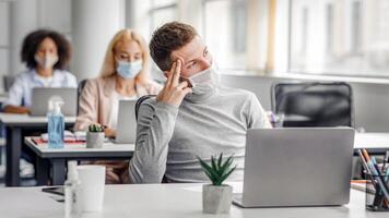 Creative ideas and brainstorming during work and social distancing. Guy in protective mask thinks and looks out the window, sitting at table with laptop with antiseptic in office interior with colleagues, empty space photo