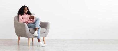 Relaxed pretty young black woman sitting in arm chair with cellphone at home, checking social media, empty white wall background, panorama with copy space. Gadgets addiction for millennials concept photo