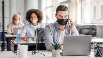 Customer consultation by phone remotely at returning to work after quarantine. Millennial man in protective mask with smartphone looks at laptop at workplace in interior of modern office with colleagues, empty space photo