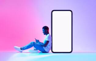 Young black man sitting next to huge smartphone with empty screen, advertising new mobile application or website, using modern gadget in neon light. Cellphone display mockup photo