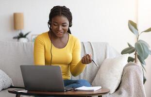 Millennial African Lady In Headset Watching Webinar On Laptop At Home, Having Online Training, Studying With Computer And Taking Notes To Notepad, Enjoying Distance Learning, Copy Space photo