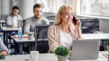Modern work with client remotely in office with technology. Smiling businesswoman talking on smartphone and looking out the window, typing on laptop at workplace in office interior with workers, empty space photo