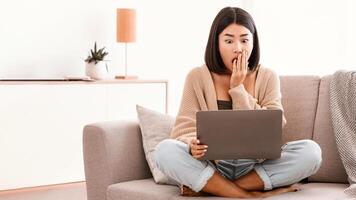Some Problem. Surprised and shocked asian lady watching something unbelievable and unexpected on computer, sitting on couch at home, copy space. Young woman with open mouth using laptop photo