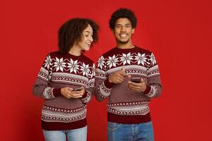 New Year and Xmas promotions and discounts in mobile app. Happy african american man points finger at smartphone and looks at camera, lady watch at his gadget, isolated on red background, studio shot photo