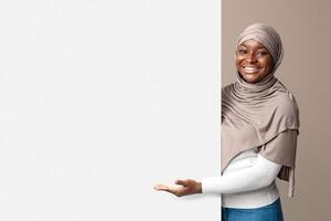 Intresting Offer. Happy Black Muslim Woman In Hijab Pointing At White Advertisement Board With Open Palm, Islamic Lady Showing Copy Space For Your Text Or Design, Standing Over Light Background photo