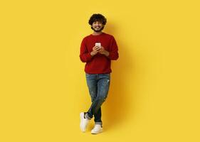 Handsome cheerful curly millennial indian guy in casual posing with cell phone in his hands on yellow studio background, smiling at camera, using nice mobile app, full length, copy space photo