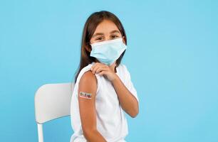 Kids Vaccination. Vaccinated Arabic Little Girl Showing Arm With Plaster After Vaccine Shot Looking At Camera Wearing Face Mask Over Blue Background. Studio Shot. Covid-19 Protection For Child photo