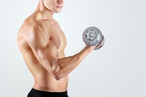Handsome Caucasian bodybuilder pumping up iron, exercising with dumbbells over light studio background. Determined millennial sportsman with bare torso working out with weights photo