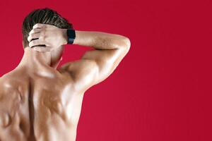 Back view of strong millennial sportsman posing over red studio background. Handsome young man with athletic sexy body demonstrating his muscles, standing with hand on his neck photo