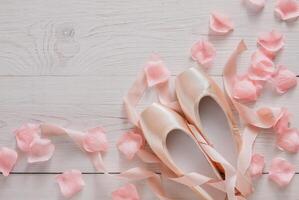 Pastel pink ballet shoes background. New pointe shoes with satin ribbon and rose petals lay on white rustic shubby chic wood, top view with copy space, soft toning photo