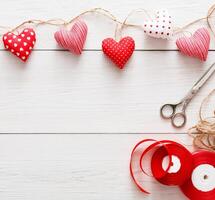 Valentine gift making, diy hobby. Sewed pillow hearts row on red clothespins border with tools for handmade at rustic white wood planks background. Happy lovers day card mockup, copy space photo