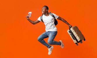 Finally vacation. Young happy black man with suitcase, backpack, passport and flight tickets jumping up orange studio background. Excited african american guy running towards copy space photo