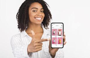 App For Online Store. Happy african woman holding and showing phone with clothes shop website and pointing at screen photo