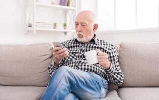 Senior man have rest using smartphone while drinking coffee or tea, sitting on sofa at home, copy space photo
