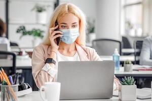 Working in modern office after returning from quarantine. Millennial lady in protective mask speaks on phone and looks at laptop at table with antiseptic in office interior, empty space photo